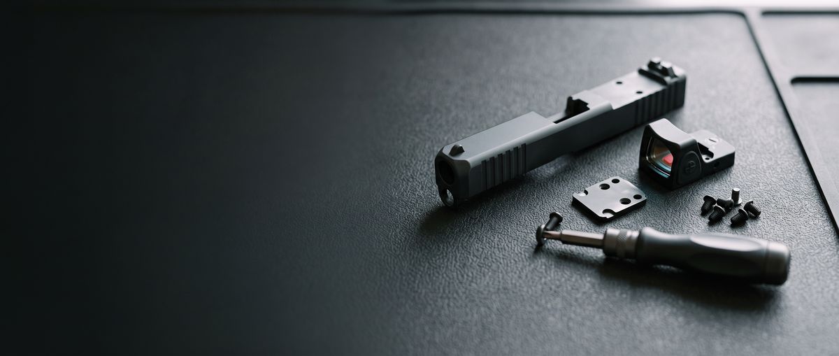 Pistol slide, red dot sight, mounting plate and screwdriver on a bench mat